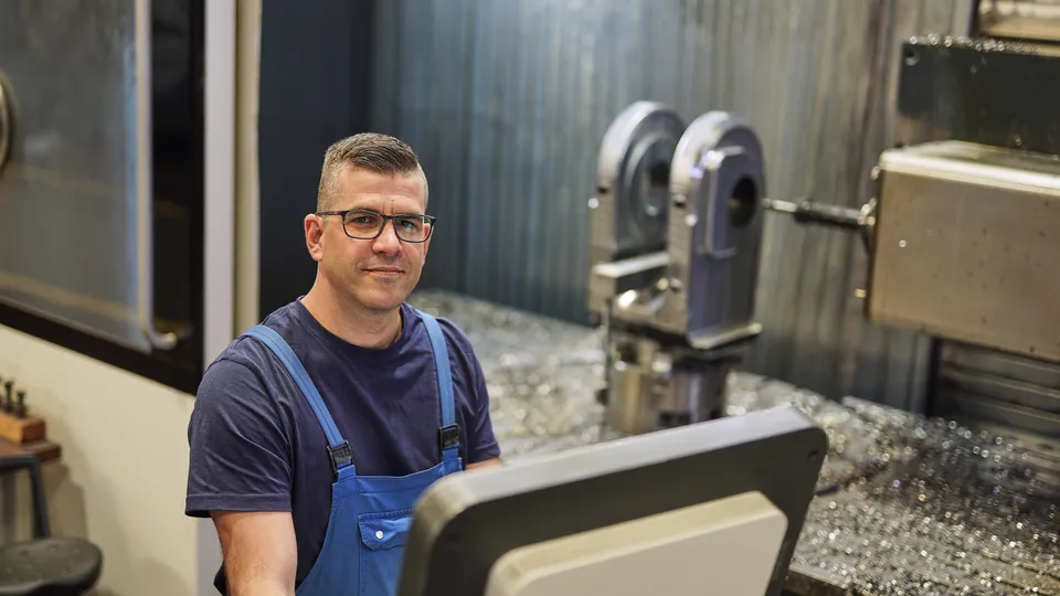 Mitarbeiter in Latzhose am Steuerpult einer CNC-Maschine blickt in die Kamera