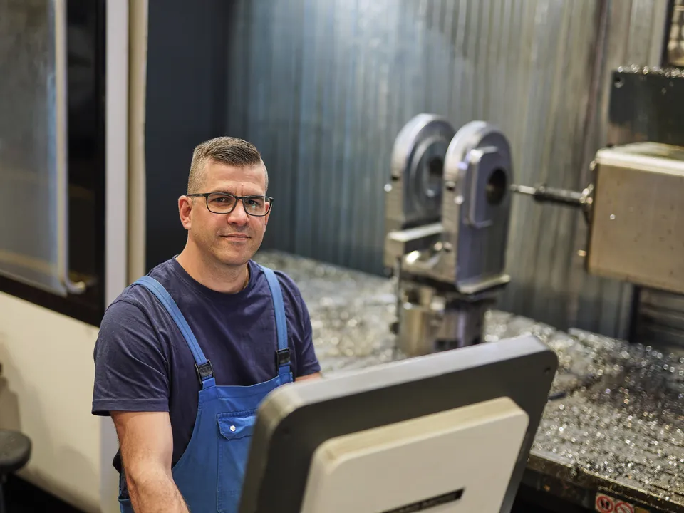 Mitarbeiter in Latzhose am Steuerpult einer CNC-Maschine blickt in die Kamera