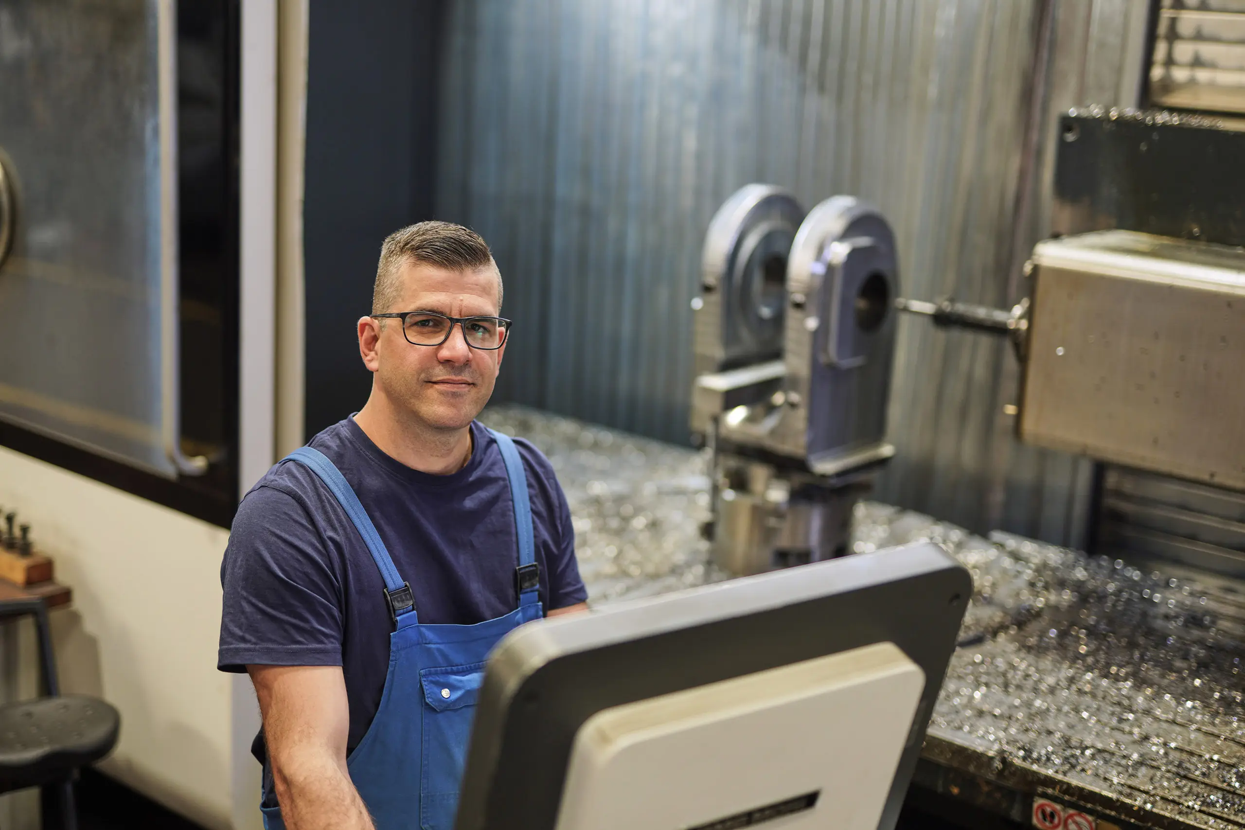 Mitarbeiter in Latzhose am Steuerpult einer CNC-Maschine blickt in die Kamera