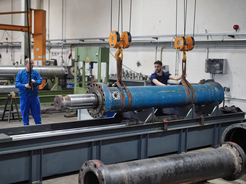 Großer blauer Hydraulikzylinder hängt am Hallenkran, zwei Mitarbeiter sichern ihn