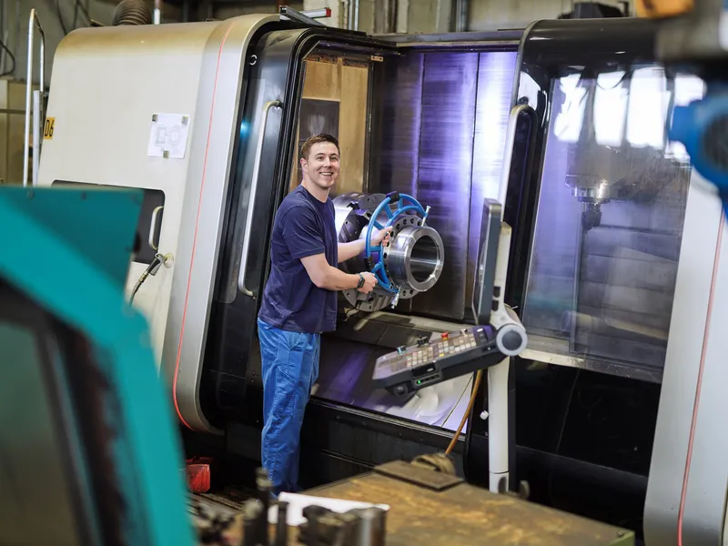 Mitarbeiter öffnet die Tür einer CNC-Maschine und hält ein gedrehtes Bauteil